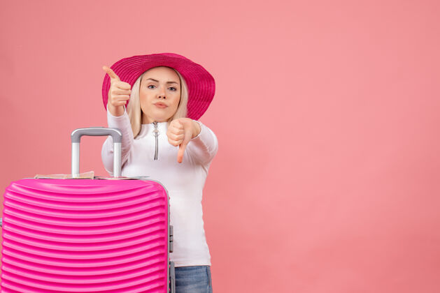前面正面图：站在粉色手提箱后面的年轻女士竖起大拇指 做着上下手势做拇指帽子