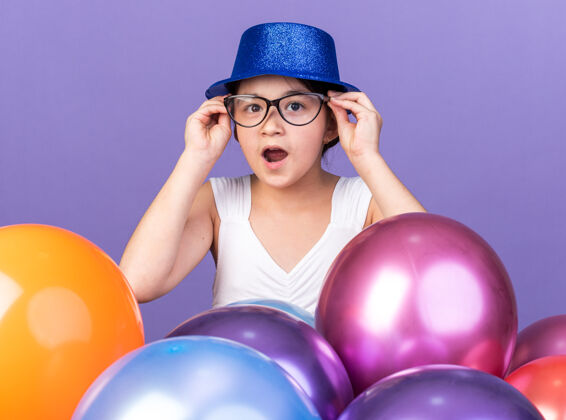 白种人惊讶的年轻白人女孩戴着蓝色的聚会帽 戴着光学眼镜 站在紫色的墙上 氦气球与复制空间隔离开来气球帽子年轻