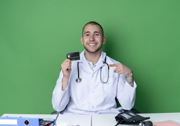 持有面带微笑的年轻男医生穿着医用长袍 听诊器坐在办公桌旁 手里拿着工作工具 指着隔离在绿墙上的信用卡穿着医生脸