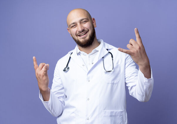 手势面带微笑的年轻秃头男医生穿着医用长袍和听诊器显示山羊的姿态年轻秃头蓝色