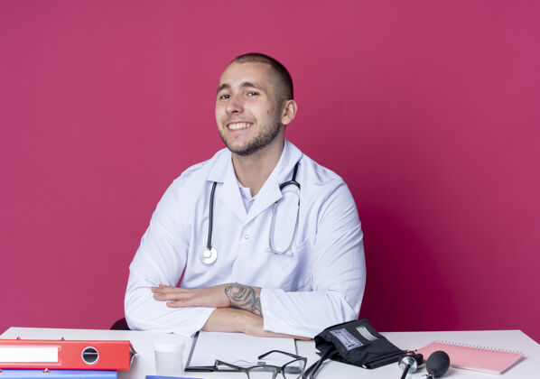 姿势面带微笑的年轻男医生穿着医用长袍和听诊器坐在办公桌旁 手放在办公桌上 隔离在粉红色的墙上医生工具年轻人