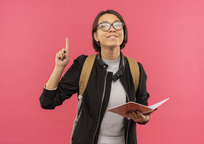 人高兴的年轻女学生戴着眼镜 背着书包拿着便笺垫 举起手指孤立在粉红色的墙上脸感觉请