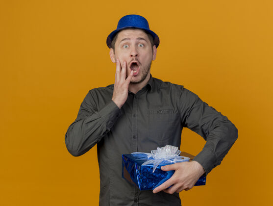 抱着害怕的年轻人戴着蓝色帽子拿着礼品盒把手放在脸颊上孤立的橙色害怕帽子派对