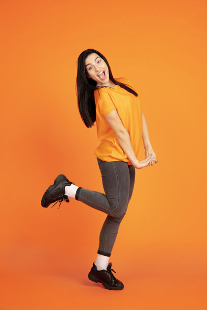年轻橙色工作室背景上的白种人年轻女子肖像穿着衬衫的漂亮黑发女性模特人类情感的概念 面部表情 销售 广告广告空间跳舞 大笑人类工作成功