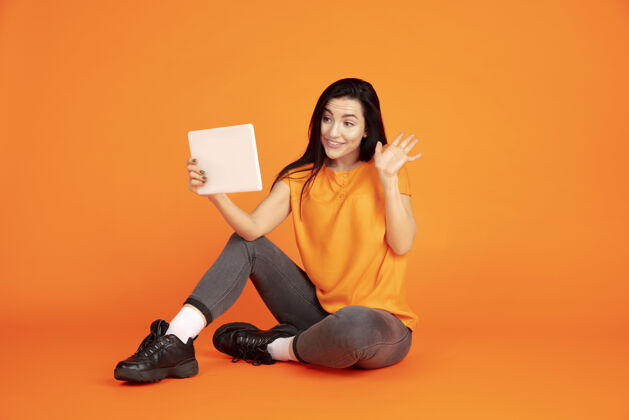 公司橙色工作室背景上的白种人年轻女子肖像穿着衬衫的漂亮黑发女性模特人类情感的概念 面部表情 销售 广告文字空间使用平板电脑 虚拟博客成功黑发人类