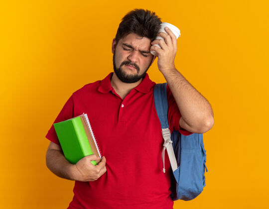 衬衫年轻的留着胡子的学生 穿着红色马球衫 背着背包 手里拿着笔记本和纸杯 站在橙色的背景下 看上去很累 而且工作过度拿着马球学生