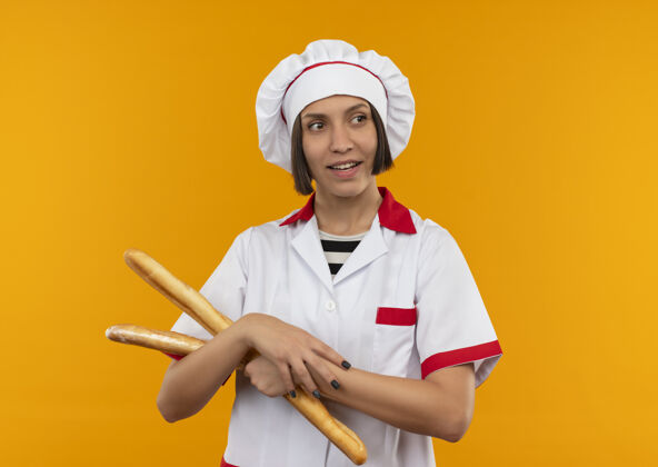 感觉微笑着的年轻女厨师身着厨师制服拿着面包棒看着隔离在橙色墙上的一面女性人握着