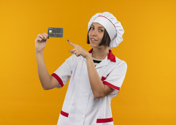 人身着厨师制服的年轻女厨师面带微笑 拿着信用卡 看着隔离在橙色墙上的门信用脸年轻人