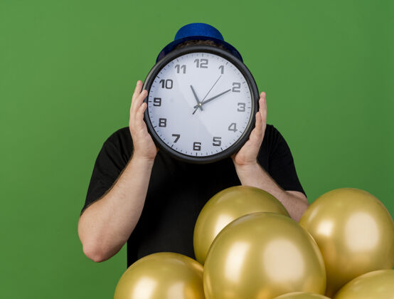 人一个戴着眼镜 戴着蓝色派对帽的焦虑的成年男子把时钟举在面前 站在绿色的墙上 氦气球被隔离开来姿势站焦虑