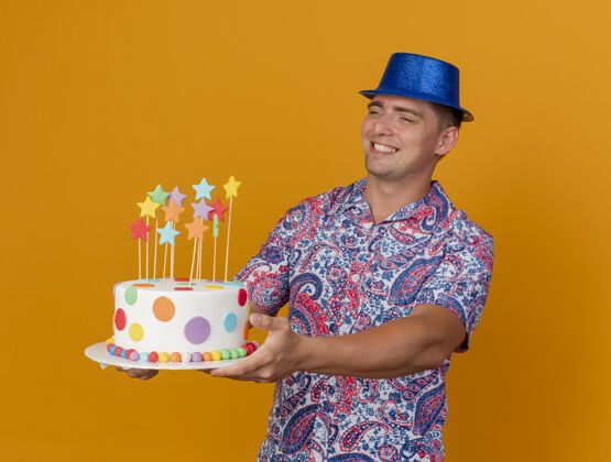 抱微笑着的年轻人戴着蓝色的帽子 拿着蛋糕站在橙色的一边派对蓝色穿