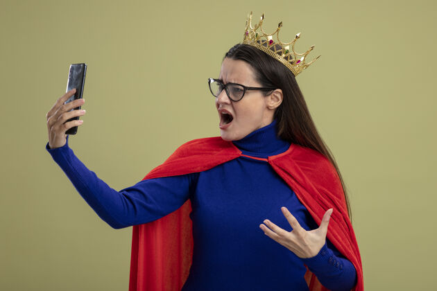 持有愤怒的年轻超级英雄女孩戴着眼镜和王冠 看着电话孤立的橄榄绿电话佩戴超级英雄