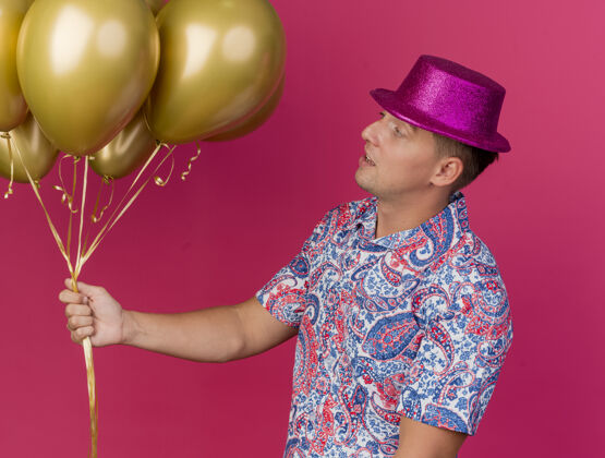 派对令人印象深刻的年轻人戴着粉红色的帽子举行聚会 看着气球孤立在粉红色戴着气球小伙子