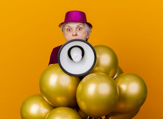 气球惊讶的老妇人戴着派对帽站着 手里拿着氦气球 把扬声器隔离在橙色的墙上女人人氦