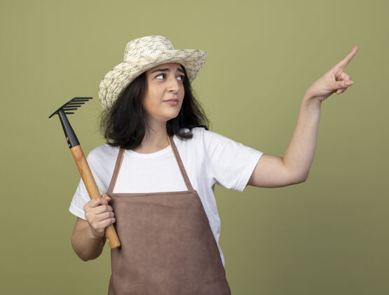 市民年轻的深色头发的女园丁穿着制服 戴着园艺帽 拿着耙子 看着橄榄绿墙上孤立的一面橄榄姿势指点