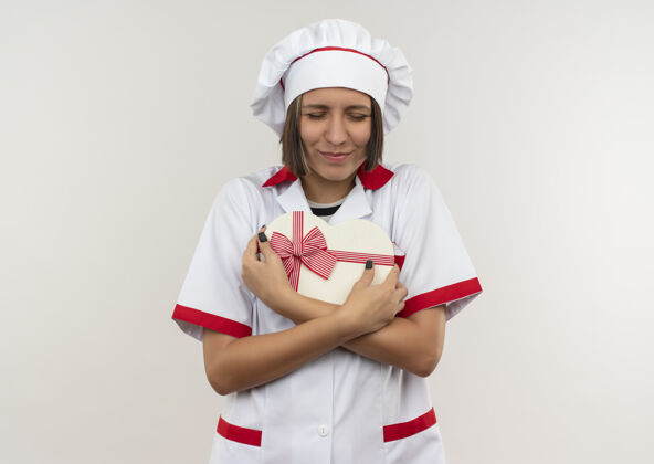 制服身着厨师制服的年轻女厨师手拿心形礼品盒 眼睛紧闭 孤零零地站在白色的墙上心形姿势感情
