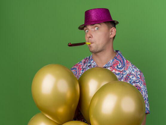 惊喜惊讶的年轻人戴着粉红色的帽子站在气球后面 吹着绿色的聚会吹风机小伙子气球派对