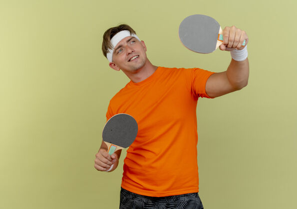乒乓球面带微笑的年轻英俊的运动型男子戴着头带和腕带举着乒乓球拍 看着橄榄绿墙上孤立的一面腕带人绿色