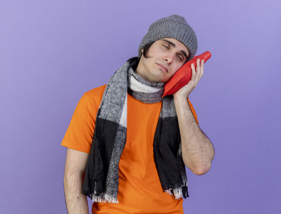 围巾歪着头的年轻病夫戴着冬天的帽子戴着围巾把热水袋放在脸颊上隔离在紫色上年轻帽子水