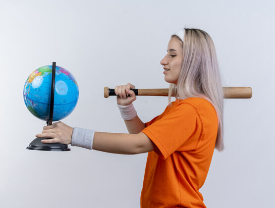 女孩戴着护腕戴着头带站在一旁拿着棒球棒和地球仪孤立地站在白墙上的年轻运动女性运动背带人