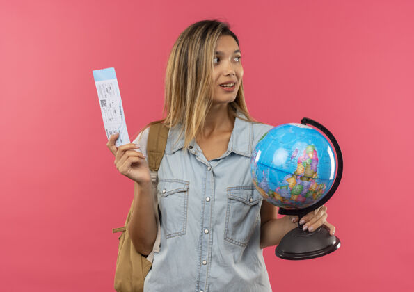 衣服高兴的年轻漂亮的学生女孩戴着背包拿着机票和地球仪看着粉红色墙上孤立的一面漂亮背人