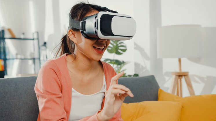 生活戴着虚拟现实耳机眼镜的亚洲女士坐在客厅的沙发上做手势可穿戴沙发房子