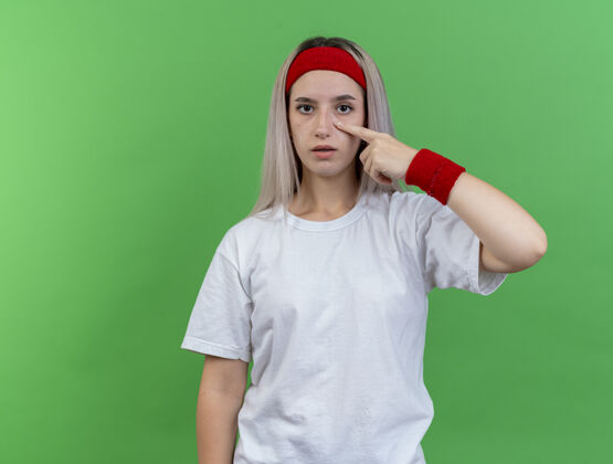 拉自信的年轻运动女性戴着背带 戴着头带和腕带 拉下眼睑 看着绿色墙壁上孤立的前方羽绒服戴人
