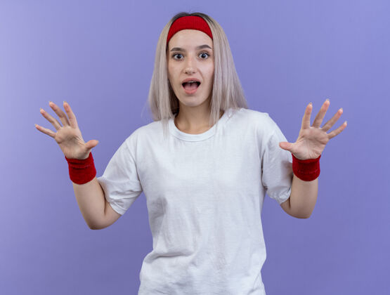 年轻惊讶的年轻运动女性戴着背带 戴着头带和腕带 在紫色的墙上张开双手背带女人运动