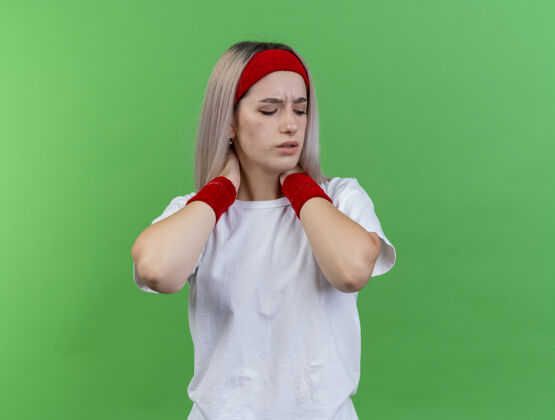 背带带着背带 戴着头带和腕带 疼痛的年轻运动女性用两只手在绿色的墙上隔离着脖子腕带女人脖子