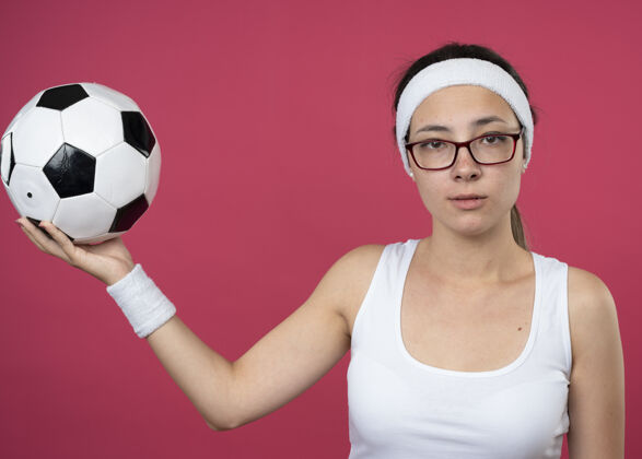 眼镜自信的年轻运动女性戴着眼镜戴着头带和腕带把球隔离在粉红色的墙上年轻人人