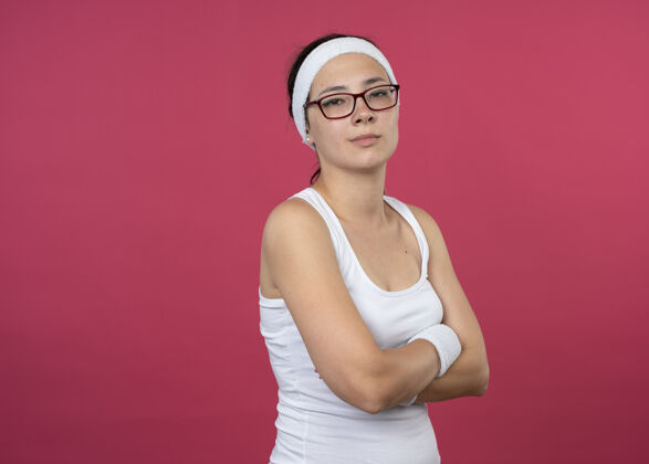 十字架自信的年轻运动女性戴着眼镜戴着头带和腕带交叉双臂站在粉红色的墙上穿光学女人