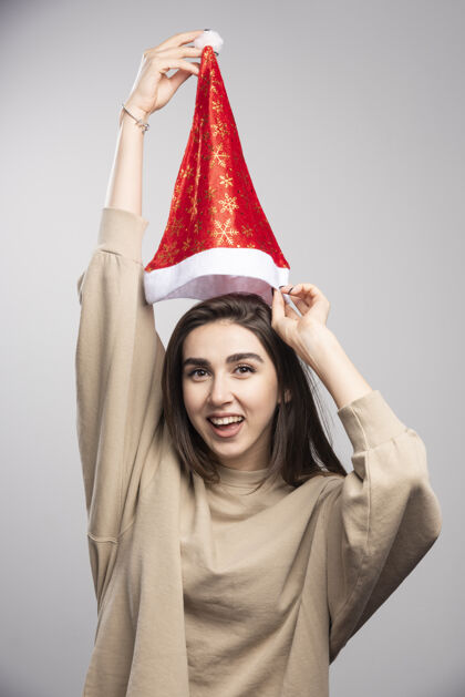 模特一个微笑的女人头顶上举着圣诞老人的帽子 背景是灰色的表情微笑女人