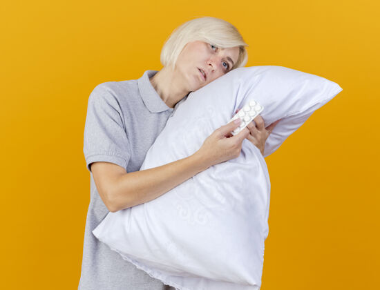 市民脸色苍白的年轻金发病女人把头靠在枕头上拿着一包隔离在橙色墙上的药丸斯拉夫表情金发