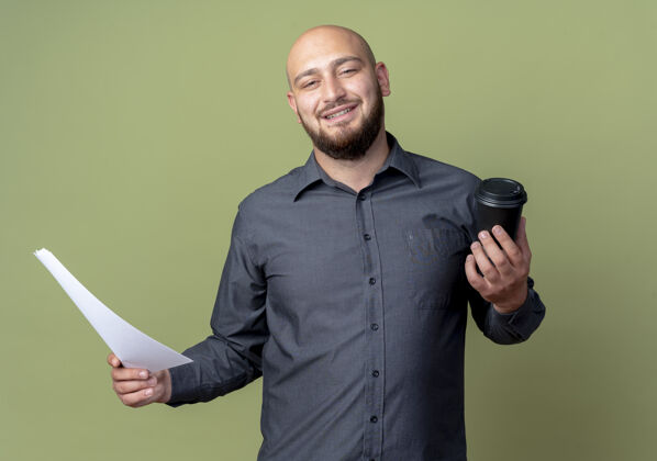 微笑微笑着的年轻秃头呼叫中心男子拿着文件和塑料咖啡杯隔离在橄榄绿的墙上杯子绿色秃头