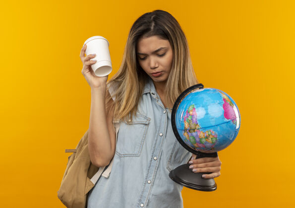 女孩悲伤的年轻漂亮的学生女孩穿着背袋拿着塑料咖啡杯和地球仪看着孤立的橙色墙上悲伤橙色公民