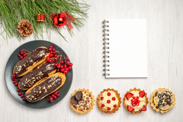 叶子顶视图：灰色盘子上的巧克力蛋糕和葡萄干底部是笔记本馅饼 白色木质地面上是松树叶子和圣诞玩具头发滑食物树