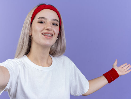 人面带微笑的年轻运动女性戴着背带 戴着头带和腕带 张开手 看着紫色墙壁上孤立的前方头带持有手