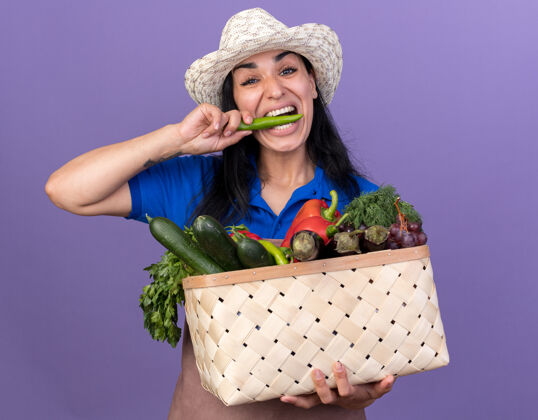 篮子年轻的白人园丁女孩穿着制服 戴着帽子 手里拿着一篮子蔬菜 看着镜头 咬着紫色背景上孤立的辣椒蔬菜女孩胡椒