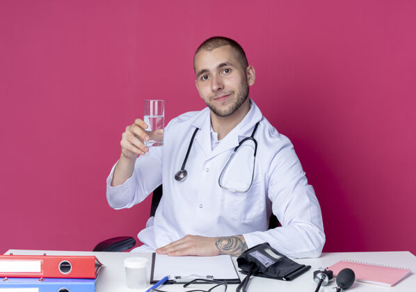 请年轻的男医生穿着医用长袍和听诊器坐在办公桌旁 手里拿着一杯水 手放在桌子上 隔离在粉红色的墙上穿着人医疗