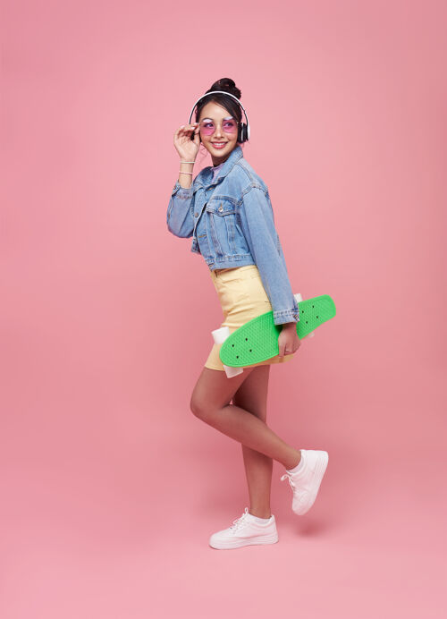 乐趣年轻的亚洲少女手持滑板 戴着无线耳机 在粉红色的墙上听音乐微笑情感快乐