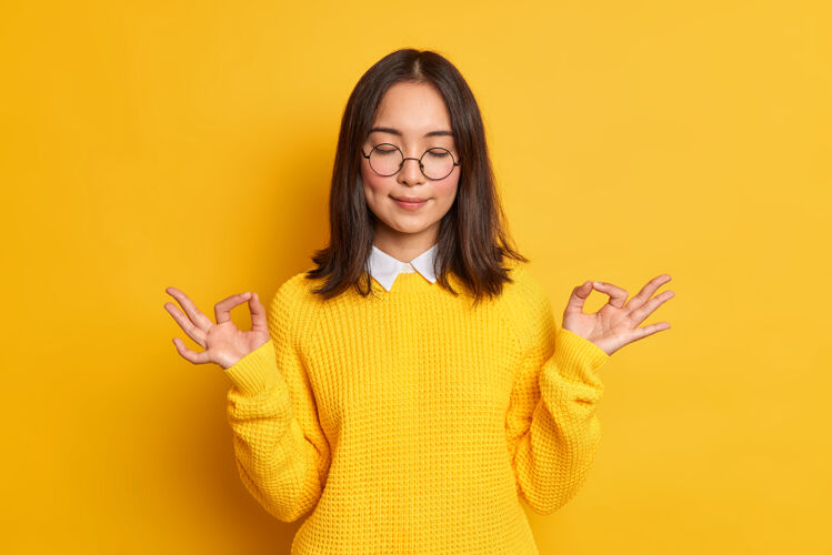 可爱冷静的亚洲年轻女子展示禅宗或OK标志冥想心情好站着闭着眼睛穿着休闲毛衣和眼镜姿势黄色眼镜