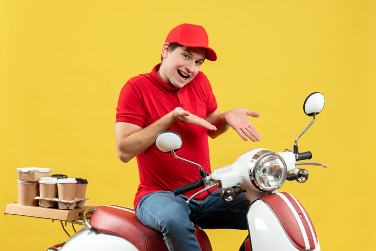 交付俯视图中微笑的年轻人穿着红色上衣 戴着帽子 指着黄色墙上的滑板车传递命令年轻人人衬衫