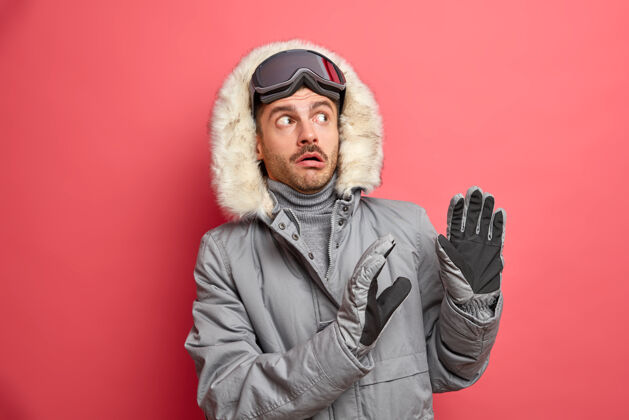 愤怒害怕的冬天男人做了一个防御的手势 害怕有什么东西要落在他身上 他穿着灰色夹克 带着皮帽和滑雪镜尴尬粉色坏