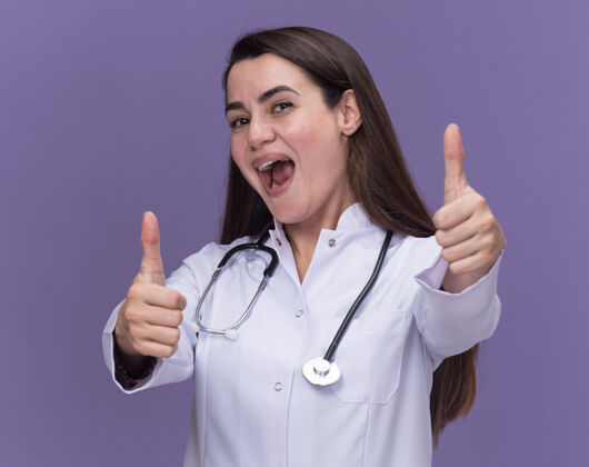 年轻快乐的年轻女医生穿着医用长袍 听诊器竖起大拇指对着紫色女听诊器长袍