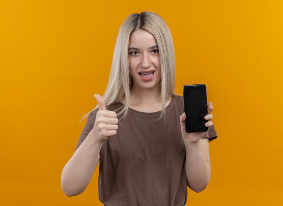 牙套戴着牙套的快乐的金发女孩拿着手机在孤立的橙色空间里竖起大拇指 还有复印空间向上快乐复制