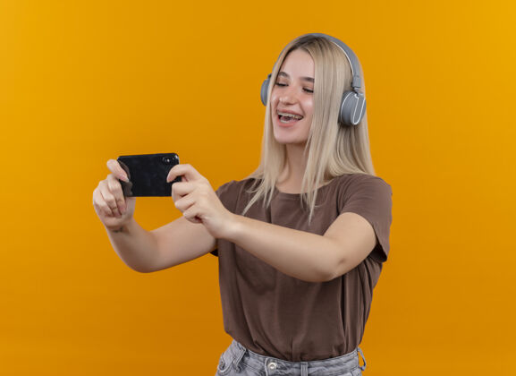 手机快乐的金发女孩戴着牙套戴着耳机拿着手机看着它在孤立的橙色空间与复印空间金发穿复制