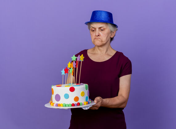 失望戴着派对帽的失望老妇人抱着生日蛋糕 看着隔离在紫色墙上的复制空间抱蛋糕老人