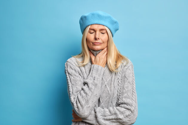 女人中年病妇摸脖子喉咙痛有流感症状闭眼止痛站在不开心的室内戴贝雷帽和暖和的针织毛衣吞咽时不愉快的感觉不适人人