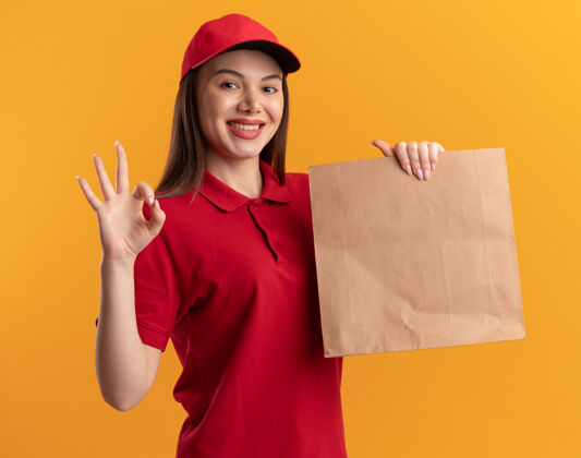 包装穿着制服的微笑漂亮的女送货员拿着纸包裹 在橙色上做着手势“ok”手势橙色手势标志
