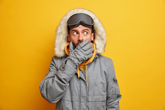 印象情绪紧张的男子滑雪板手捂着嘴试图隐藏秘密 戴着温暖的手套和灰色保暖夹克黄色害怕拼图