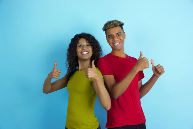 笑竖起大拇指 面带微笑年轻感性的非裔美国人男女 穿着蓝色背景的彩色衣服美丽的情侣人类情感的概念 面部表情 关系 广告 友谊化妆人指向